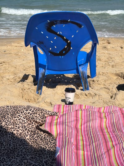 The image depicts a beach scene in Ashkelon, Israel. In the foreground, two beach towels are spread on the sand: one with a leopard print and another with vibrant pink, green, and purple stripes. A blue plastic chair with a spray-painted black "S" is positioned behind the towels, partially embedded in the sand. Next to the chair, a disposable coffee cup with the "Corona Extra" logo is visible. In the background, the calm turquoise water of the Mediterranean Sea meets the sandy shore. The overall impression is one of a relaxed day at the beach.