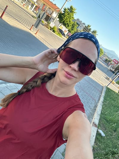 A woman with blonde hair in a braid and wearing a red tank top and sunglasses is standing on a sidewalk in Fethiye, Turkey. She is holding her hair with her right hand and looking at the camera. The background shows a street with buildings, trees, and mountains. There are also traffic cones and a sign for "BIM". The sun is shining and the sky is blue.