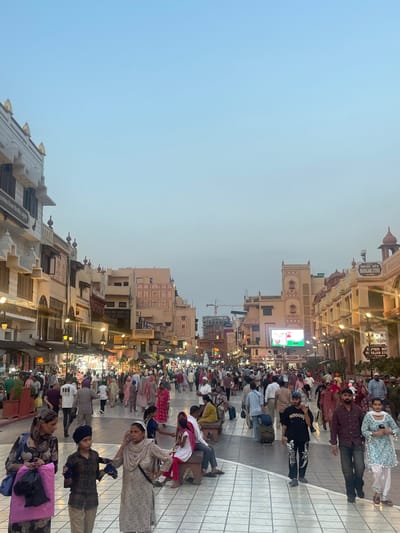 The image depicts a bustling street scene in Amritsar, India, likely near the Golden Temple. The street is paved with patterned tiles and lined with buildings showcasing traditional Indian architecture with decorative facades and balconies. Numerous people are walking in both directions, many dressed in traditional Indian attire. Some are carrying shopping bags, while others are pushing luggage. Market stalls are visible along the sides of the street, displaying various goods. Streetlights illuminate the area, suggesting it is either early morning or late afternoon. A large electronic billboard is visible in the distance. The overall atmosphere is lively and dynamic, indicative of a popular public space.