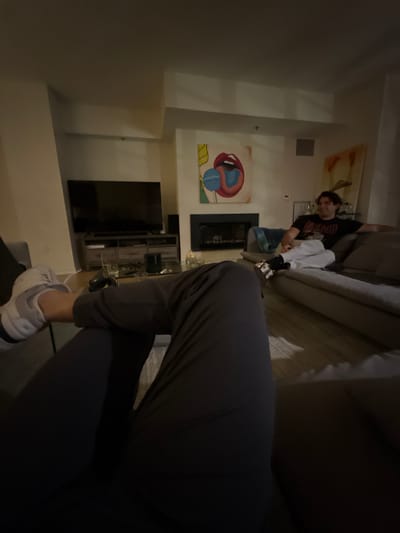 A man is on a sofa, partially visible, wearing a black t-shirt with a design. In the foreground, a person wearing gray athletic pants and white sneakers is seen. A television is mounted on the wall, beneath a painting featuring an abstract, colorful representation of lips and a sphere. A fireplace is situated between the artwork and the television. A cabinet with decorative items is placed below the television.
