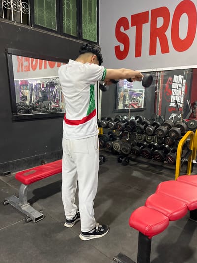 The image displays an indoor gymnasium located in Fez, Morocco. A male individual is depicted from the rear, standing upright with his back to the camera, performing a lateral raise exercise using a black dumbbell in each hand, held at shoulder height with arms extended horizontally. He is dressed in a white polo shirt featuring green and red striped accents on the left side and sleeve, white ribbed athletic pants, and black and white New Balance sneakers. His dark, curly hair is styled with faded sides.
The gym environment includes a dark grey wall with large mirrored sections. These mirrors reflect various gym equipment, including racks of dumbbells and other exercise machines in the background. Reversed text, including "STRONGER" and "TRAIN LIKE THE BEAST," is visible in the mirror reflections. Above the mirrored section on the right, the partial red lettering "STRO" is painted directly on the wall. A yellow metal rack filled with numerous black and silver dumbbells is situated behind the exercising individual. Several additional dumbbells are scattered on the dark rubber gym floor. Two red-padded workout benches are present: a flat bench to the left and a multi-section adjustable bench partially visible on the right. A window with white security bars is observable above the left mirrored section.