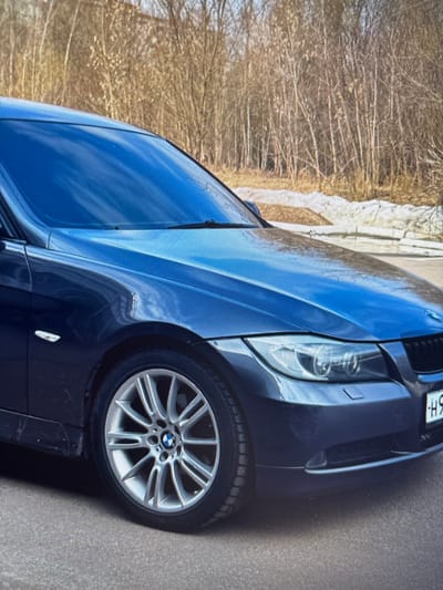 A dark blue BMW sedan is parked on an asphalt road. The car has a dent on the front passenger side fender. The background shows a line of bare trees and some patchy snow on the ground, suggesting a cold-weather environment. The vehicle's front right wheel and part of the front bumper are visible. The license plate is partially obscured but shows Cyrillic characters. The scene appears to be outdoors, possibly in a suburban or park area, given the natural elements in the background. No people are present in the image. The lighting suggests it might be daytime.