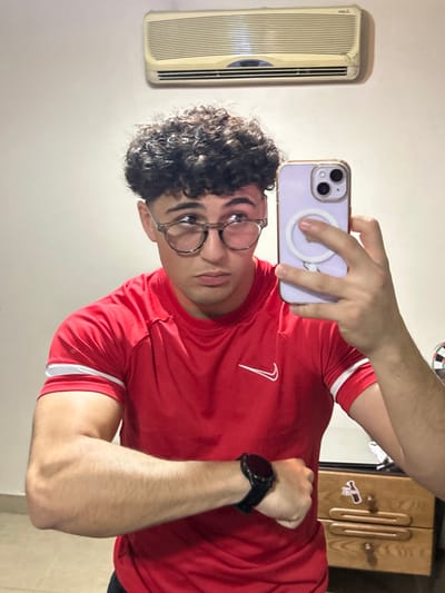 The image displays a young male individual reflected in a mirror, capturing a selfie indoors. He possesses dark, curly hair and wears round, light-colored eyeglasses. His attire consists of a red short-sleeved athletic shirt with a white Nike swoosh logo on the chest and two white stripes on the right sleeve. A black smartwatch is present on his left wrist, which is flexed in the frame. His left hand holds a light purple smartphone, equipped with a transparent case and a white circular MagSafe accessory, directed at the mirror. The background visible in the reflection features a plain white wall, an off-white wall-mounted air conditioning unit with horizontal louvers in the upper section, and partially visible on the right, a wooden dresser with two drawers and a portion of a dartboard. The image itself contains no specific visual indicators to confirm a location within Sarouja Municipality, Syria.