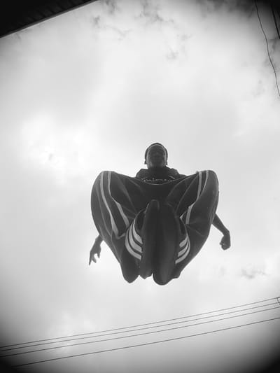A low-angle, black and white, medium shot captures a Black male in mid-air, performing a jump or athletic maneuver. He is positioned centrally in the frame, his legs bent and drawn upwards, creating a tucked or seated pose. He wears dark track pants with white stripes down the sides and a dark t-shirt. His gaze is directed towards the camera. The background consists of an overcast sky with visible clouds and overhead power lines, suggesting an outdoor urban environment. The perspective is from below, looking up at the subject against the sky. The image appears to have been taken in Narayi, Nigeria, given the context.