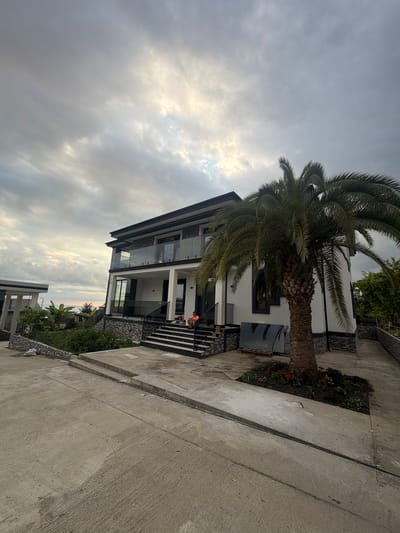 An exterior, wide-angle, eye-level shot captures a modern, two-story white house with dark trim and glass balconies. The house is situated on a slight incline, with a concrete pathway leading up to a set of dark-tiled stairs. A large palm tree stands prominently in front of the house, its fronds reaching towards the overcast sky. To the left of the house, another modern structure with white columns and a flat roof is partially visible. Lush green vegetation and some flowers are planted around the base of the palm tree and in front of the structures. A person wearing an orange shirt is seated on the stairs. The sky is filled with dramatic, textured clouds. The scene appears to be in a residential area, possibly near a coast given the presence of palm trees and the distant, hazy view of water. The overall impression is one of a newly constructed or renovated property.