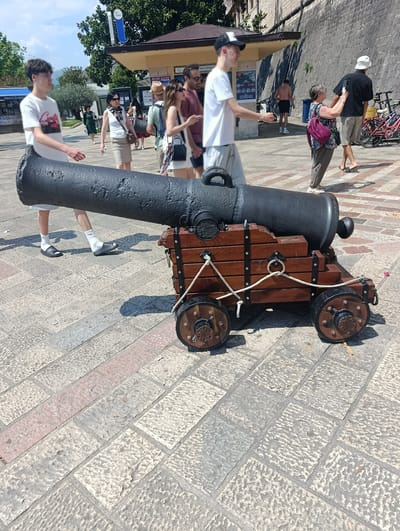 In Kotor, Montenegro, a life-sized replica of a cannon on a wooden carriage is positioned on a stone-paved walkway. The cannon barrel is black and metallic, with a detailed wooden carriage featuring two wheels on each side. The foreground is dominated by the cannon, with its wheels and carriage clearly visible.

In the background, several people are walking and interacting. A young man in a white t-shirt and light-colored pants stands to the left of the cannon, looking towards the right. Behind him, a group of people are walking away from the camera. These include a woman in a white top and a tan skirt, a man in a straw hat, a woman in a white top with sunglasses, and a man in a dark t-shirt and shorts. Further in the background, a man in a white hat and a dark shirt walks away, accompanied by a woman in a patterned top and a purple backpack. Several bicycles are parked on the right side of the frame.

The environment appears to be a sunny day with a blue sky. Trees are visible in the background, along with a building that has a sign that reads "INFORMA" and "RENT A BIKE". The walkway is made of interlocking stone pavers in various shades of grey and pinkish-red. The overall scene captures a moment of daily life in a historic setting.