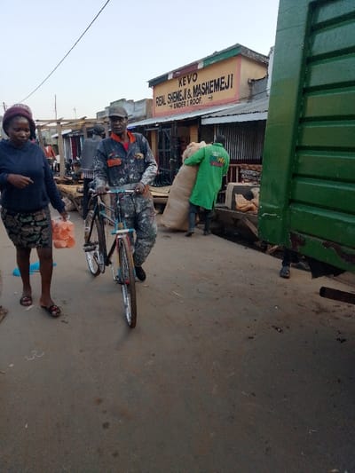 The image shows a street in Malava, Kenya (latitude: 0.489, longitude: 34.837). A man in a blue and camouflage jumpsuit is riding a bicycle in the street,  while a woman in a black and floral patterned dress walks to his left. The man is carrying a stack of orange plastic cups in his right hand. A second man in a green jacket is walking in the opposite direction carrying a large sack. There are buildings along the street, with one bearing the sign "KEVO REAL SHEMEJI & MASHEMEJI UNDER 1 ROOF." A large, green truck is parked to the right, partially obscuring the scene. The street is paved and the ground is dusty.  The sky is clear and blue.  The lighting is bright, suggesting midday.
