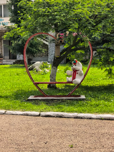 In Sterlitamak, Russia, a white teddy bear wearing a red scarf and a multicolored hat sits on a red heart-shaped swing in a grassy area near a building. The swing is supported by a metal frame. There are trees with green leaves and a birdhouse hanging from a branch. The ground in front of the swing is covered in green grass, and a paved path is visible in the foreground. The building in the background appears to be made of stone and has windows.