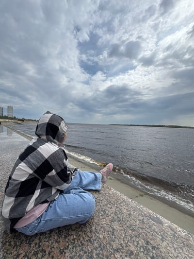 A person wearing blue jeans, pink sneakers, and a black and white checkered hooded shirt sits on a granite barrier next to a body of water under a cloudy sky. The person faces the water. The photo appears to be taken near a riverbank in Nizhnevartovsk, Russia, based on the skyline visible in the distance. Waves from the water lap against the concrete barrier.