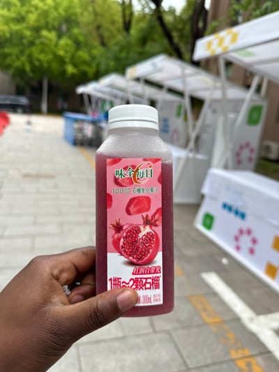 A close-up, slightly rotated shot shows a dark-skinned hand holding a clear plastic bottle of pomegranate juice. The bottle has a white screw cap and contains a red-purple liquid. Its label features a vibrant design with images of fresh pomegranates, both whole and halved, showcasing their arils.

The visible Chinese text on the label, according to OCR, includes the brand name "味全每日" (Wei Chuan Daily), and product description "100% 石榴复合果汁" (100% Pomegranate Compound Juice). Marketing phrases like "红润自带光" (Rosy and Luminous) and "1瓶~2颗石榴" (1 bottle ~ 2 pomegranates) are also present, indicating the juice contains the equivalent of two pomegranates. The volume is specified as "300mL", with a disclaimer "图片仅供参考" (Images for reference only).

The background is an out-of-focus outdoor setting, suggesting a street or pathway. Blurred green trees are visible, along with white structures that appear to be part of a display or booth. The ground below shows light-colored concrete with faint yellow markings. The lighting suggests it is daytime, possibly overcast, given the lack of harsh shadows. The Chinese text on the bottle aligns with the given context of Daxing District, China.