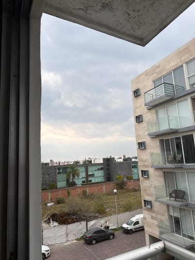 The image depicts an upward view from a window, likely in San Andrés Cholula, Mexico, characterized by urban development and partly cloudy skies. Two multi-story buildings are visible, one with a modern facade featuring balconies, large windows, and neutral-toned stone cladding, and another with a mix of glass and darker materials. A landscaped area is present between the buildings, featuring grass, bushes, and a few trees, and a chain-link fence. A street with parked cars is to the side. The foreground features the frame of the window, which is made of metal. No people are clearly identifiable.