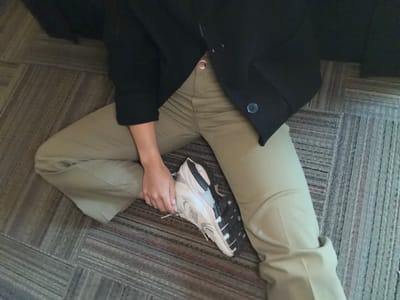A person is sitting on a carpeted floor. They are wearing khaki pants and a black blazer. A white and gray New Balance sneaker is near their left leg, held by their left hand. The location cannot be determined.