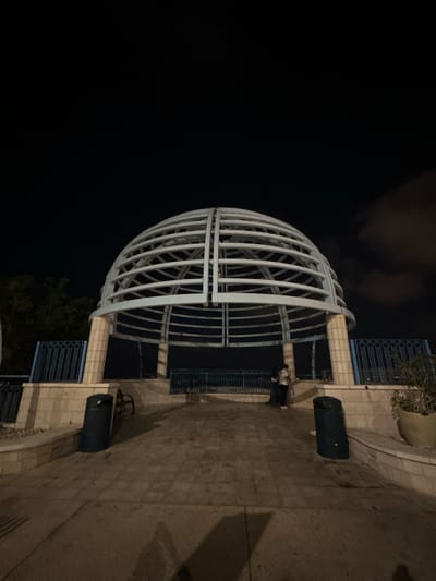 The image captures a night scene of a domed, open-air structure located in the city of Haifa, Israel. The structure, made of white metal ribs, sits atop a tiered stone base. In front of the structure, a paved pathway leads towards it, with two dark green trash receptacles visible on either side. A metal railing with vertical blue bars lines the perimeter of the elevated area. To the left, trees are visible against the dark sky, and to the right, a large potted plant is situated. Two individuals, dressed casually, are standing near the entrance of the domed structure, facing away from the camera. The sky is dark, with some clouds visible on the right side of the frame. The lighting suggests artificial illumination from the immediate vicinity.