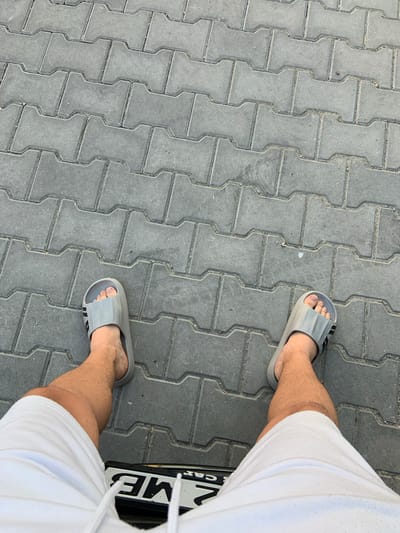 A first-person perspective shot shows a person's legs and feet standing on a gray interlocking paver sidewalk. The person is wearing white shorts and gray slide sandals with black stripes. A license plate is visible beneath the person's legs, partially obscured by the shorts. The license plate has black characters on a white background and appears to be from Bulgaria. The overall scene is a common urban or suburban setting, possibly in the city of Byala, Bulgaria, given the license plate.
