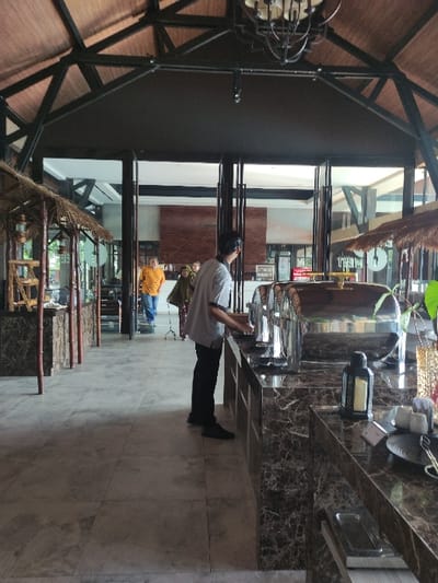 The image shows a man in a white shirt and black pants standing on a tiled floor in a restaurant in Gambiran, Indonesia (latitude: -8.434, longitude: 114.18). He is looking down at a large food warmer with a silver dome. The restaurant has a modern, minimalist design with wooden beams and large windows. There are other people sitting at a table in the background. The image is captured from a low angle, looking up at the man from the floor. The scene is a typical restaurant setting, with a focus on food preparation and service.