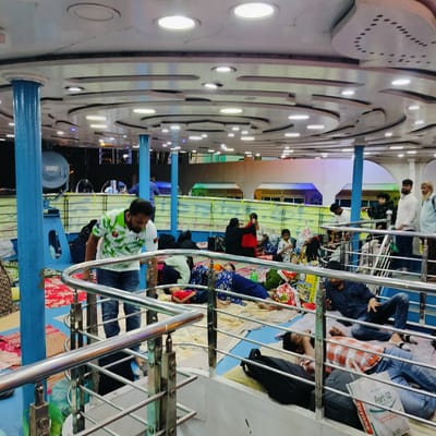 The image captures the crowded interior of a passenger ferry, likely at night, indicated by the sole reliance on artificial lighting. The ceiling features a multi-tiered, decorative design with numerous circular recessed lights providing illumination. Large, blue cylindrical pillars support the structure, complemented by chrome-plated railings that define walkways and different levels of the deck.
Many people are visible throughout the space, primarily resting or sleeping on various colorful rugs and mats spread across the blue-painted floor. Their personal belongings, including backpacks and other bags, are scattered beside them. In the foreground, a man wearing a green and white patterned shirt and blue jeans walks down a step past the railings. Below him, a man in a red and white plaid shirt and jeans is lying down, resting, with a black backpack and a white shopping bag beside him. Another man in a dark blue shirt is sitting further back, engrossed in his phone.
Further into the scene, groups of individuals, including women and children, are seated or lying down. One woman in black is seen adjusting a red garment. In the background, several men are standing, observing the surroundings. The distant background shows windows or open sections, revealing indistinct exterior structures with colorful lighting.
Visible text includes "AVIAZ" in green and "SQURE" in red on the white shopping bag in the foreground. A yellow banner with indistinct script, possibly Bengali, stretches across a partition in the middle ground. The scene depicts a bustling environment on a mode of transport, where people are engaged in travel and rest, consistent with a ferry service in Bhola, Bangladesh.