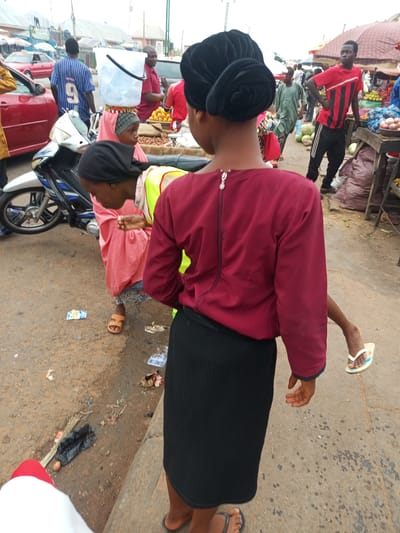 In Tempure, Nigeria, this image captures a bustling street market scene. The foreground is dominated by the back of a young woman wearing a maroon long-sleeved top and a black skirt. Her dark hair is styled in a bun. To her left, a child in a pink garment is partially visible, holding something in their hand. Behind them, a person is carrying a large white bucket on their head. The background reveals various market stalls with produce, people in colorful attire, and vehicles, including a red car and a motorcycle. The ground is dusty and littered with debris, indicative of a busy urban environment.