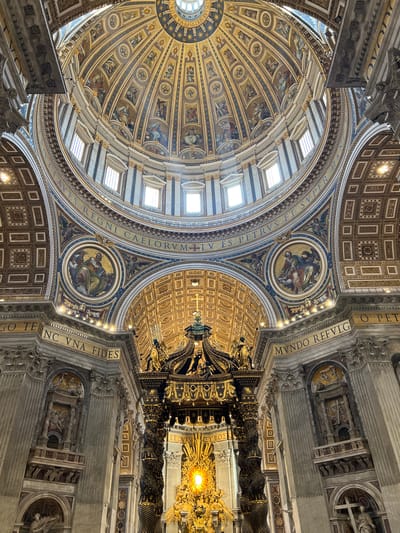 This is an upward-angled, wide interior shot of St. Peter's Basilica in Città del Vaticano, Vatican City, showcasing its grand dome, the Bernini Baldachin, and the Cathedra Petri.
The central focus is the monumental, gilded bronze Bernini Baldachin, featuring four massive twisted columns with ornate detailing and a crown-like canopy, positioned over what appears to be the papal altar. Behind and above the baldachin, the radiant golden Cathedra Petri (Chair of St. Peter) is visible, glowing brightly from an internal light source, crowned by a representation of the Holy Spirit as a dove within a burst of golden rays.
Dominating the upper half of the image is the massive dome of St. Peter's Basilica. Its interior is exquisitely decorated with gilded coffering and intricate panels depicting numerous religious figures, primarily saints or apostles, arranged in radial segments. The drum of the dome features several rectangular windows, allowing natural light to illuminate the space.
Below the dome's drum, at the level of the pendentives, two large circular mosaic medallions are prominently visible. The left medallion depicts a robed figure seated, holding a book, with a bull symbol (traditionally St. Luke). The right medallion shows a similar figure with an eagle symbol (traditionally St. John the Evangelist). Surrounding these medallions and encircling the base of the dome, a Latin inscription in golden letters on a blue background is visible, stating "TV ES PETRVS ET SVPER HANC PETRAM AEDIFICABO ECCLESIAM MEAM ET TIBI DABO CLAVES REGNI CAELORVM" (Thou art Peter, and upon this rock I will build my church, and I will give unto thee the keys of the kingdom of heaven).
The surrounding architectural elements include massive fluted columns or pilasters, with highly decorated capitals and entablature. In the background, on either side of the baldachin, large niches are carved into the walls. The left niche features multiple levels of sculptures, while the right niche contains a large cross and a figure below it. The ceilings of the surrounding structures are also richly coffered and gilded. The color palette is dominated by gold, deep blues, white, and various tones of marble, creating an opulent and awe-inspiring atmosphere. No discernible people are present in the image.