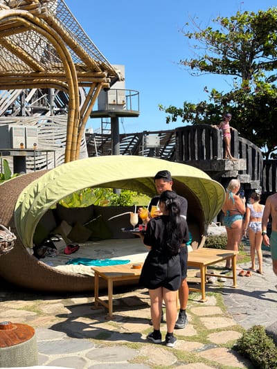This is a vertical, eye-level, outdoor shot on a sunny day. The scene appears to be a resort or a cafe with a pool area, possibly in Kediri, Indonesia. In the foreground, a circular, woven daybed with a light green canopy is prominent. Two individuals are seated on the daybed, facing away from the camera. The person on the left, a man, is wearing a black t-shirt and dark shorts. The person on the right, a woman, has long black hair and is wearing a black top and black shorts. She is holding two drinks with yellow and orange liquid, one with a straw and a garnish.
Behind the daybed, a wooden chair is positioned. The ground is paved with large, irregular stones interspersed with grass. In the mid-ground, there is a black, multi-tiered structure that appears to be a staircase or a decorative element, possibly with water features. A person in pink shorts and a pink top is visible on this structure, leaning down. Other individuals in swimwear are also visible in the background, some sitting and one taking a photo with a phone.
To the left of the frame, a large tree with lush green foliage extends from above, casting shadows on the scene. In the far background, there is a glimpse of a body of water, likely a swimming pool, and more resort structures, including a tall bamboo-like construction. The time of day is suggested to be midday due to the strong sunlight and clear blue sky. The overall mood is relaxed and recreational.