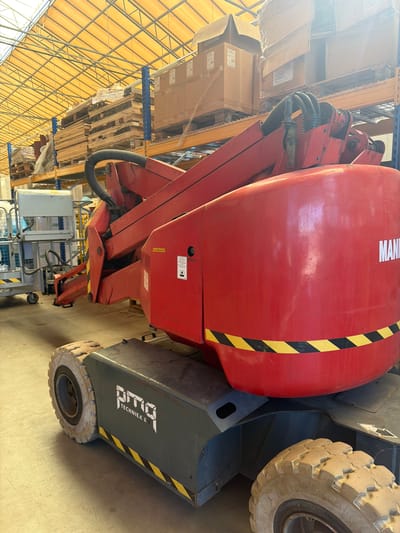 The media file shows a large red articulated boom lift parked indoors, likely in a warehouse or industrial setting. The boom lift is the primary subject, with its large red boom prominently featured. The texture of the red paint is visible, with some scuffs and wear. A black and yellow striped safety marking runs diagonally across the boom. The word "MANITOU" is visible in white lettering on the boom.
To the right of the red boom is the dark gray chassis of the lift, including a large, rough-treaded tire. The text "RIGO TECHNIEK B" is visible on the side of the chassis in white lettering, with a similar black and yellow striped safety marking below it.
In the background, shelves stacked with boxes and wooden pallets are visible. The shelving units are made of blue metal, and the overall impression is of a storage or distribution facility. The lighting suggests it is daytime, with a warm, artificial light source. The concrete floor is visible beneath the machine. The scene is static, indicating the lift is not in operation at the moment. The location context is Maasdijk, Netherlands.