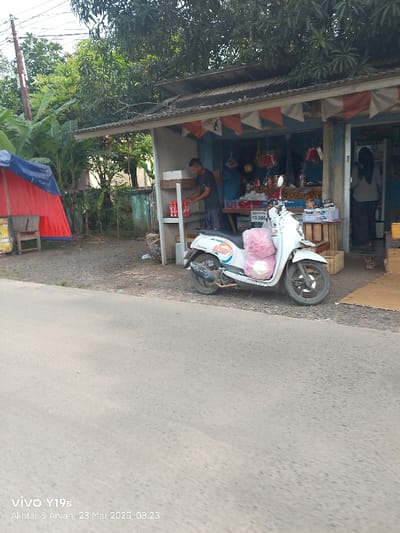 The scene is a street in Kecamatan Anjatan, Indonesia (latitude: -6.418, longitude: 107.965) on a sunny day. A man in a blue shirt and jeans is standing in front of a small shop, reaching for a bottle on a shelf. He is facing the right side of the image. A woman in a black hijab is visible inside the shop. A white motorcycle is parked on the street in front of the shop. There are pink bags on the seat of the motorcycle. There are also some items and goods on the shelf of the shop. The shop has a light blue painted exterior. The street is paved with asphalt. The shop has a sign that says "Buket Bunga 10.000". There are trees and a blue tarpaulin tent visible in the background. There is a small bench behind the tent. The scene is taken from the street, looking towards the shop. The image has a timestamp of 23 Mar 2023, 08.23. There are power lines visible above the shop. There are some goods and items in the shop, including red drinks, and some fruits. There is also a red cooler bag visible.