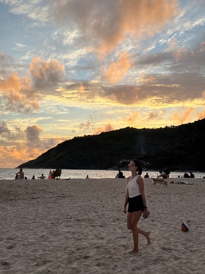 The image depicts a sunset on a sandy beach in Rawai, Thailand. The sky is filled with dramatic clouds painted in hues of orange, pink, and blue, suggesting the golden hour. The sun is setting behind a dark, vegetated hill that slopes down to the ocean, creating a striking silhouette. The calm sea reflects the colorful sky.
On the beach, several people are visible, engaged in various activities. In the foreground, a woman in a white tank top and black shorts is standing, looking down at something she's holding, possibly a phone or a small container. Her posture suggests a moment of relaxation or contemplation. Further down the beach, there are other individuals and groups, some appearing to be walking, others sitting or standing. A volleyball is visible in the sand near the woman in the foreground, hinting at recreational activities. A few dogs are also present on the beach, some interacting with people. The overall atmosphere appears to be peaceful and leisurely, characteristic of an evening at the beach. The sand is textured with small ripples from the receding tide.