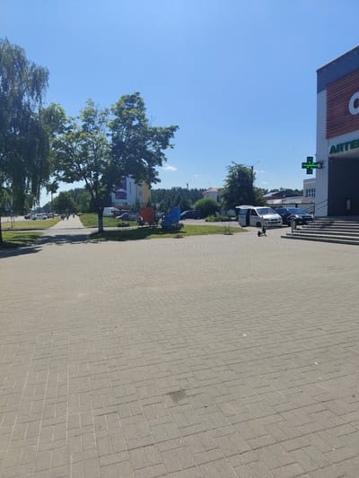 The image depicts a paved walkway in front of a modern building, likely a commercial establishment, in Salihorsk, Belarus. The building features a large "C" logo and signage that appears to be "АПТЕКА" (Pharmacy). A green cross symbol, commonly associated with pharmacies, is also visible on the building's exterior. In the foreground, the walkway is paved with rectangular grey bricks. To the right of the walkway, a set of stairs leads up to the building's entrance. Several vehicles are parked in the vicinity, including a white minivan with its doors open and a dark-colored car. A black electric scooter is positioned on the walkway near the minivan. Beyond the immediate area, a line of trees forms a backdrop, with a hint of other buildings and the horizon visible in the distance. The sky is clear and blue, indicating a sunny day. The scene appears to be a typical urban streetscape.