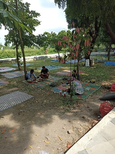 This image captures an outdoor gathering in Mataram, Indonesia, with several people enjoying a meal in a relaxed, natural setting. The scene is set on a grassy area under the partial shade of trees, indicating a bright, sunny day, likely during midday or early afternoon due to the distinct shadows.
The main subjects are a group of individuals, predominantly men, sitting on patterned woven mats and blue tarps spread across the grass. In the foreground, two men are seated facing each other on a mat, actively eating from plates laid before them. Various food items, dishes, and bottles of condiments are visible on their mat. Further back and to the right, another person, partly obscured by a small tree with distinctive reddish-pink leaves, is also seated on a mat, engaged in eating. Several other mats, some occupied and some empty, are scattered across the grassy area, suggesting a larger communal event or multiple small groups.
The setting includes mature trees providing shade, and the ground is a mix of grass and exposed dirt with fallen leaves. In the background, a low concrete barrier separates the picnic area from what appears to be an expansive green field, possibly rice paddies, under a bright sky with some clouds. Bags are visible near some trees and mats, possibly containing personal belongings or trash. The overall atmosphere appears casual and social, characteristic of an outdoor meal or picnic. There is no visible text in the image.