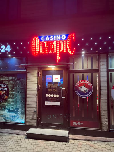 This image captures the exterior facade of a commercial establishment, identified as an "Olympic Casino," located in Liepaja, Latvia. The scene is illuminated by artificial lighting, suggesting nighttime or low-light conditions.
The central feature is a large, vibrant neon sign spelling "CASINO" in blue letters on a curved backdrop and "OLYMPIC" in prominent red, dotted capital letters. To the left of "CASINO," a partially visible blue neon "24" indicates 24-hour operation. The facade above and around the main sign also features smaller, evenly spaced white lights.
The entrance consists of a dark-framed glass door. Affixed to the door are several informational signs and stickers: "SPORTA BĀRS UN KAZINO" (Sports Bar and Casino) with operating hours "00-24" (24 hours), and an age restriction statement "liela no 18 gadu vecuma" (persons over 18 years of age). Below this, a row of red circular icons with crossed-out symbols denotes prohibited activities or items, alongside a QR code. An "OlyBet" brand logo is also visible on the door.
To the left of the entrance door, a large window displays a backlit graphic depicting a city skyline at night, with additional images including beverages. To the right of the door, an adjacent glass panel features a large circular sticker with a red, white, and blue design, displaying an image of individuals and the text "SPORTA TIESRAIDES" (Sports Broadcasts). The building's facade surrounding the glass sections is constructed from vertical wooden or wood-paneled sections, which appear weathered in some areas.
Directly in front of the entrance door, a low, rectangular dark-colored step or platform is present. The ground level is paved with interlocking cobblestones. No individuals, vehicles, or observable actions or interactions are depicted in the visible frame of the image.