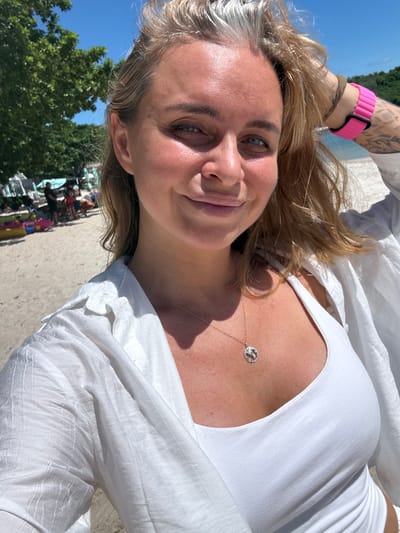 A woman takes a selfie on a sunny beach. She is wearing a white tank top and a white open shirt. She has blonde hair and is wearing a silver necklace with a round pendant. On her left wrist, she is wearing a bright pink watch. In the background, there are trees, beach chairs, and people. The location is Choeng Mon beach in Thailand.