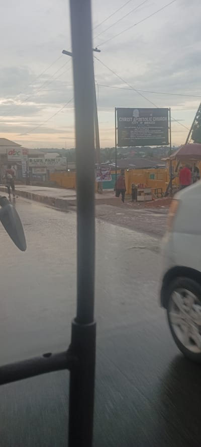 The image shows a street scene in Umuahia, Nigeria, likely captured from a moving vehicle. The wet road suggests recent rain. Prominent landmarks include a sign for the "Christ Apostolic Church (City of Miracles)" and storefronts advertising "B-LUX PAINTS NIG. LTD. DEPOT" and "abc". Several pedestrians are visible on the sidewalks, and a parked white vehicle is on the right side of the road. The foreground is partially obscured by part of the vehicle the picture was taken from, including a mirror and frame element. Overhead power lines are also present.