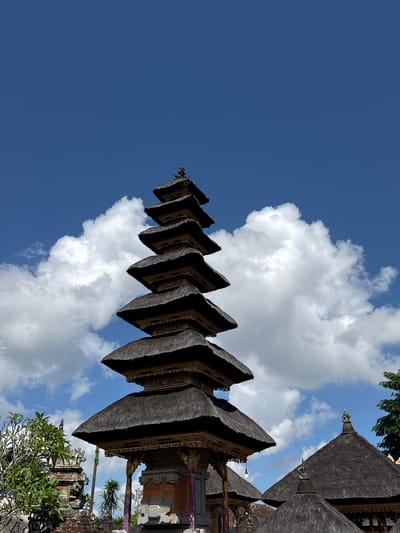 The image captures a tiered pagoda-style temple, likely a Meru tower, situated in a tropical environment. The structure features multiple levels with thatched roofs, each progressively smaller as they ascend. The pagoda is adorned with intricate golden embellishments and is set against a vibrant blue sky dotted with fluffy white clouds. In the foreground, to the left, lush green foliage and another temple structure with stone carvings are visible, suggesting a temple complex. To the right and in the background, more thatched-roof structures and trees can be seen. The overall scene conveys a sense of spiritual and cultural significance within a serene natural setting, characteristic of Ubud District, Indonesia. There are no people or animals visible in the image.