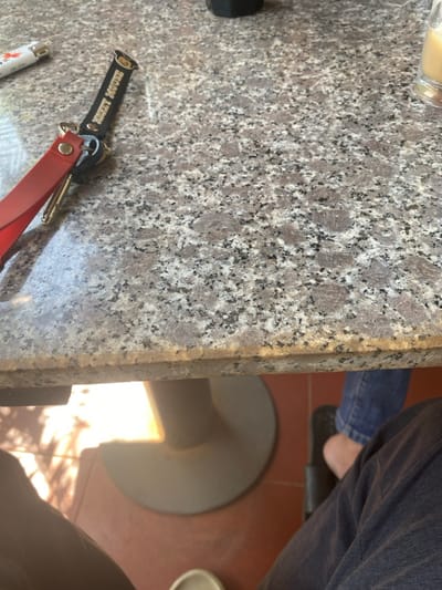 The scene depicts a granular granite-patterned tabletop, light grey with black and brown speckles, situated in Đak Đoa, Vietnam. On the upper left of the table rests a white lighter with orange markings and a set of keys. The keys are attached to a red leather strap and a black keychain bearing the text "MICKEY MOUSE" in yellow, accompanied by a yellow Mickey Mouse head graphic. A dark, cylindrical object is partially visible at the upper center. In the upper right corner, the rim of a clear glass containing a light brown liquid is visible. Below the table, a light-colored, flared cylindrical table base is centered on a reddish-brown tiled floor. Direct sunlight illuminates a section of the floor, creating defined shadows. The lower legs of two individuals are visible; one in the lower left wearing dark pants and light footwear, and another in the lower right wearing blue jeans and a dark sandal. Both individuals are seated at the table.