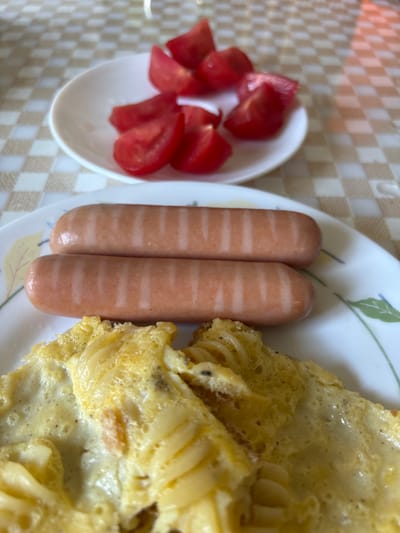 A close-up, slightly angled shot shows a breakfast plate with two sausages, an omelet, and a small dish of sliced tomatoes. The sausages are pink and have faint, lighter stripes. The omelet is golden yellow and appears fluffy, with some darker specks, possibly pepper, visible. The sliced tomatoes are bright red and wedge-shaped. The food is arranged on a white plate with a subtle floral design. The plate rests on a checkered tablecloth with alternating beige and white squares. The background is slightly out of focus, showing more of the tablecloth. There is no indication of people, actions, or interactions, and no specific location in Bila Tserkva, Ukraine, is identifiable.