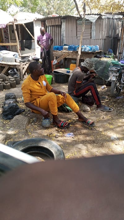 This image depicts an outdoor scene in Fagge A, Nigeria, characterized by informal market activity or a communal gathering area under bright daylight.
In the foreground, an adult male is seated on the dry dirt ground, facing right and slightly upward. He wears a mustard yellow long-sleeved tunic and matching trousers, with black sandals featuring red and green "GUCCI" branding on his feet. A small, light-blue, decorated ceramic pot with a handle rests in his left hand. A folded white item is visible in his left shirt pocket. To his left, several dark, log-like wooden pieces are stacked, alongside black plastic bags on the ground. A discarded vehicle tire is partially visible in the immediate foreground.
To the right of the first man, another adult male is seated on the ground, wearing a dark brown shirt with orange accents, dark trousers with red stripes, and a patterned bucket hat. He is focused downwards, manipulating an object in his hands, possibly a fishing net or fabric. He wears grey slip-on sandals. Behind him, a silver motorcycle is partially visible.
In the background, an adult male stands, wearing a pink t-shirt and dark trousers, holding dark fabric or clothing. He is positioned near a rudimentary wooden stall with an open frame. Behind him, several structures constructed from corrugated metal sheets and wood are visible. A wooden table in the mid-background displays numerous blue-wrapped items, possibly sachets or small bags of products, arranged in rows. Other objects include various plastic buckets (green and black), a silver kettle, and scattered debris on the dirt ground, which is covered with dry leaves and small stones. Sparse trees and dry foliage are visible in the upper background. The scene is illuminated by strong sunlight, casting shadows.