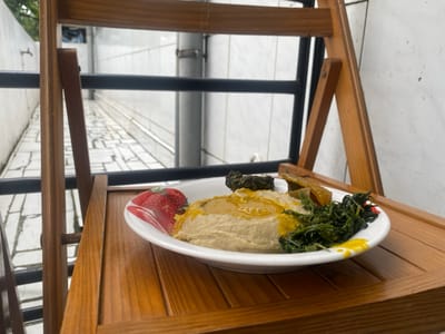 A plate of food sits on a wooden table. The plate contains a mound of light brown mashed food, topped with a yellow sauce and a small amount of green vegetables. There are also some other pieces of food on the plate, including a darker green vegetable and some yellow, possibly fried, pieces. The food is located on a wooden folding chair, which is positioned in front of a wall. The wall is white, but the texture appears to be either painted or plastered. There is a black metal railing behind the chair, and the railing is made of horizontal bars. There is a narrow, white walkway behind the railing, lined with white stone blocks. The walkway leads into a building, which is barely visible in the background. There are two small green plants visible on the top of the wall, and another green plant is barely visible in the upper left-hand corner of the image. The coordinates provided (latitude: 4.007, longitude: 9.211) correspond to a location in the city of Limbe, Cameroon.