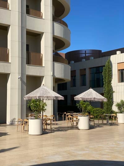 This is a medium-shot, outdoor photograph taken on a sunny day, depicting an outdoor seating area with two decorative umbrellas and surrounding vegetation. The scene is situated in front of a modern building with a curved architectural design and balconies.
In the foreground, a light-colored tiled patio is visible, with long shadows cast by the umbrellas and surrounding objects, indicating the sun is to the left and slightly behind the viewer. Two large, white cylindrical planters are prominent. The left planter contains a leafy green plant and is shaded by a white, macrame-style umbrella with a fringed edge. The right planter also contains a similar green plant and is under a second, identical umbrella. Around these planters, several light-wood chairs with woven seats surround small, round tables.
Behind the seating area, more greenery and tall cypress trees are visible. The background features a multi-story building with a cream-colored facade, accented by dark-framed windows and gold-colored balcony railings. Further back, another building with a darker, curved wooden facade and windows can be seen.
The image appears to have been taken on the grounds of the Sirius Park hotel in Sochi, Russia, a location known for its contemporary architecture and landscaped outdoor spaces. There are no people visible in the photograph.