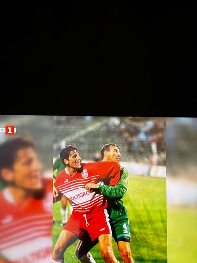 The image displays a split view of two soccer players engaged in a match. On the left, a player in a blurred red and white striped jersey is partially visible, with a portion of a logo and the text "MULTIGRO" visible on his shirt. To the right, a player in a red jersey with white stripes and a crest is in focus, with the word "MULTIGRO" also visible on his shirt. He is grappling with another player in a green jersey. The background appears to be a soccer field with spectators in the distance. The split image may be from a broadcast or a visual editing technique. The presence of the logo and text suggests a sponsored event or team. The scene is indicative of a professional or amateur soccer game. There is no specific information provided in the image that would place it in Ognianovo, Bulgaria.