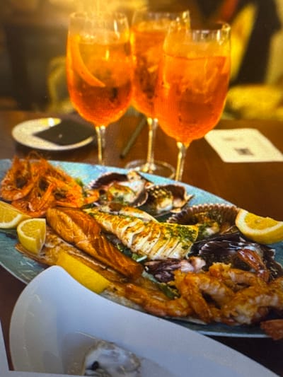 Three glasses of orange aperitif are in the background, along with a plate of seafood including prawns, scallops, and fish, garnished with lemon wedges. The table setting appears to be in a restaurant. No people are visible in the image. The location in the city of Murcia, Spain, is unknown.