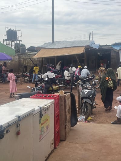 The image depicts a street scene in Lafia, Nigeria, likely a market or a busy thoroughfare, on an overcast day. In the foreground, a white chest freezer is visible, secured with a padlock, next to red crates. Beyond the freezer, a busy street unfolds with numerous motorcycles, some parked and others with riders. People are seen interacting and moving around. A woman in a pink dress is walking away from the viewer, and further back, a woman in a dark hijab with a baby strapped to her back is walking towards the right of the frame. Several men are present, some on motorcycles, and others standing or walking. Structures with corrugated metal roofs are visible in the background, characteristic of informal settlements or marketplaces. A water tank on a metal platform is also noticeable in the distance. The ground appears to be dirt or gravel. The overall impression is one of daily life and commerce in a Nigerian urban setting.