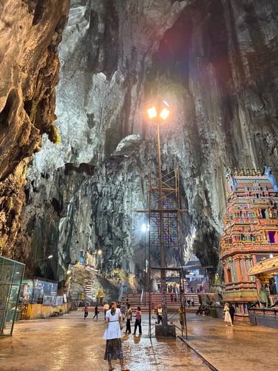 This media file captures the interior of a vast, naturally formed cave, likely a temple complex. The scene is dominated by towering, rugged cave walls and a high ceiling, illuminated by numerous artificial lights, including bright floodlights on poles. The ground is a wide, flat, and wet-looking concrete surface, reflecting the light.

In the mid-ground, high up on a cave ledge, stands an intricately decorated Hindu temple structure, possibly a gopuram, adorned with vibrant colors like red, orange, blue, and gold, and detailed carvings. Below this temple, scaffolding or temporary structures are visible. Further into the cave, a series of white stairs with red trim and railings ascend, indicating multiple levels for visitors.

Several people are present, walking on the wet floor or ascending the stairs. One woman in the foreground wears a white top and a leopard print skirt, walking towards the right. Others appear further back on the stairs, suggesting a pilgrimage or tourist activity. The overall impression is one of awe-inspiring natural geology combined with significant religious architecture.

Notable details include the dramatic scale of the cave, the contrast between the natural rock formations and the colorful man-made structures, and the bright artificial lighting creating a vivid atmosphere. The wet ground suggests either natural water seepage or recent cleaning. Visible text on a green barrier reads "Pusat Rekreasi & Kebudayaan" (Recreation & Culture Center), reinforcing the site's function. The setting is consistent with the Batu Caves in Selayang Municipal Council, Malaysia.