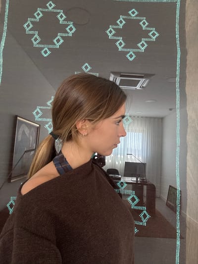 The image shows a person with brown hair, wearing a brown sweater and a patterned neck covering. They are indoors, possibly in a room with a decorative rug. The rug is patterned with geometric designs. The background shows a glimpse of an interior room setting with a window, desk, and artwork. The image does not directly indicate the location being Torres Vedras, Portugal.