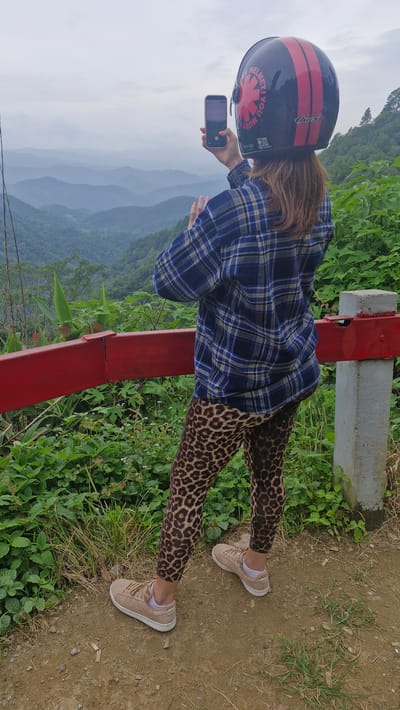 In Samoeng Tai, Thailand, a person stands on a dirt path overlooking a valley of lush green mountains under a cloudy sky. The individual, facing away from the camera, is wearing a blue and yellow plaid long-sleeved shirt, leopard-print leggings, and beige sneakers with white socks. They have a black motorcycle helmet with red stripes and a red chili pepper logo on their head, and are holding a smartphone in their right hand, appearing to take a photo or video. A red metal railing runs horizontally in the foreground, partially obscuring the person's lower legs. Green foliage, including tall grasses and leafy plants, grows around the railing and the path. A concrete post supports the railing on the right side of the frame.