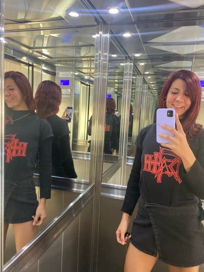 This image captures a single female subject taking a selfie inside an elevator, identified as being in Wilde, Argentina. The subject, positioned centrally, has reddish-brown hair styled shoulder-length and is smiling, revealing teeth. She wears a black long-sleeved top underneath a black short-sleeved t-shirt featuring a prominent red, stylized "DEATH" logo on the chest. She is also wearing a black mini-skirt and a thin silver necklace. Her right hand holds a mobile phone, encased in a light purple protective cover, pointed towards herself to capture the image. Her left hand holds a small black spherical object.

The elevator interior is characterized by highly reflective mirrored walls and polished metal panels, which create numerous reflections of the subject, extending infinitely into the background. The ceiling is also reflective, featuring several illuminated circular recessed lights. A metallic handrail is visible along the side walls. In the reflections, a digital display showing a blue numeral "0" and a metallic plaque with partially visible Spanish text, including "CAPACIDAD PERSONAS" (Capacity Persons), are discernible. The floor appears to be a dark, possibly metallic, material.