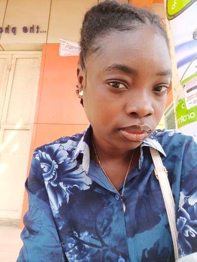 A medium-shot portrait depicts a dark-skinned individual, identified as a young adult female, looking directly at the camera with a neutral expression. Her dark, textured hair is styled back. She wears a dark blue collared shirt featuring a lighter blue floral pattern and a small, gold-colored heart-shaped stud earring in her visible right ear. A thin gold-colored chain necklace with a curved, light-colored pendant is visible, along with the light-colored strap of a shoulder bag crossing her right shoulder. The background to the left features an orange-colored wall segment positioned above a light-colored, diamond-patterned metal shutter or door. Above the orange wall, an inverted purple sign partially displays text such as "tdq eht...". To the right, a vertical pole or column is partially covered with promotional posters, including a prominent green section with white text, where "orio" is discernible. The scene is located in the city of Fagge A, Nigeria.