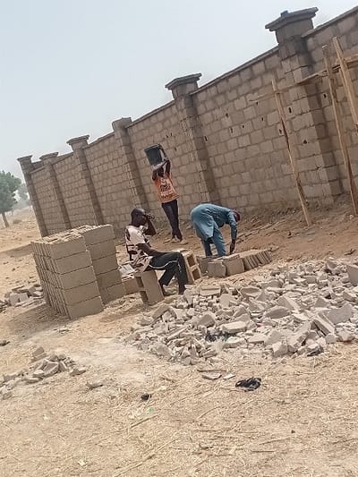 The image displays an outdoor construction scene in Ayama, Nigeria. Three adult males are present near a partially constructed perimeter wall made of light grey concrete blocks. The wall extends across the mid-ground and features multiple vertical pillar sections topped with decorative capstones.
In the left foreground, a male individual is seated on a stack of unlaid concrete blocks. He is wearing a light-colored, patterned shirt and dark trousers. His right arm is bent and resting.
In the center-right, another male, dressed in an orange t-shirt with visible text "500" and dark trousers, is standing and holding a dark bucket with both hands, lifted above his head, suggesting material transport.
To the far right, a third male, clad in a light blue long-sleeved garment, is bent over, engaged in ground-level work, possibly arranging or handling construction materials.
A large pile of broken concrete blocks and rubble occupies the central foreground. A distinct stack of whole concrete blocks is located in the lower-left. The ground is dry, light brown earth with sparse dry vegetation. In the far right, two rudimentary wooden poles, possibly temporary supports, stand vertically. The background on the left shows distant green trees under a hazy, bright sky.