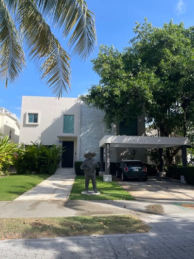 The image shows a modern two-story house with a white exterior and gray stone accents, located in Playa del Carmen, Mexico. The house has a green front door and a covered carport with a dark green car parked underneath. A statue of a man in a sombrero stands on the lawn in front of the house. Lush tropical foliage surrounds the property, including palm trees visible in the foreground. The sky is clear and blue. A paved walkway leads to the front door, and a concrete driveway extends to the street. Another house is partially visible on the left of the frame.