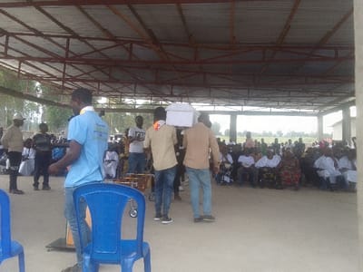 A group of people is gathered under a metal roof structure. A crowd of seated individuals occupies the right side of the frame. Several men stand near the center and left. Two men carry a white object, possibly a box, on their shoulders. A man in a blue shirt stands in the foreground. Blue plastic chairs are scattered around, and trees are visible in the background.