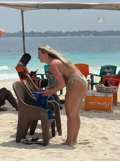 A sunny beach scene unfolds in Zanzibar, Tanzania, featuring white sand, clear turquoise ocean water, and a distant green landmass under a pale sky. In the foreground, a fair-skinned female with long blonde hair, wearing a light green crocheted two-piece bikini, is bending forward towards three brown plastic chairs. She has a gold bracelet on her right wrist, and her bare feet are in the sand. Blue fabric is draped over one of the brown chairs. To her right, an orange cooler with the word "TUMBI" partially visible in white lettering supports a tray holding numerous small clear plastic bags filled with snacks.
In the mid-ground, a dark-skinned male, wearing an orange and black striped short-sleeved shirt and black shorts, is seated on an orange plastic chair, looking down at a smartphone. Behind him, a green plastic chair holds a grey bag. Further to the left, only the legs of another dark-skinned individual in dark shorts are visible, seated on an unseen chair. A large grey beach umbrella provides shade over part of the scene. In the upper right background, a white bird is captured mid-flight against the sky and ocean. Waves gently break near the shore.