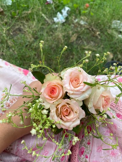 In Tarabo, Bangladesh, a person's forearm rests on a pink fabric with a floral pattern, holding a small bouquet of pale pink roses and delicate white wildflowers. The background consists of green grass with some scattered debris, suggesting an outdoor setting, possibly a park or garden. The lighting appears to be natural daylight, casting soft shadows.
